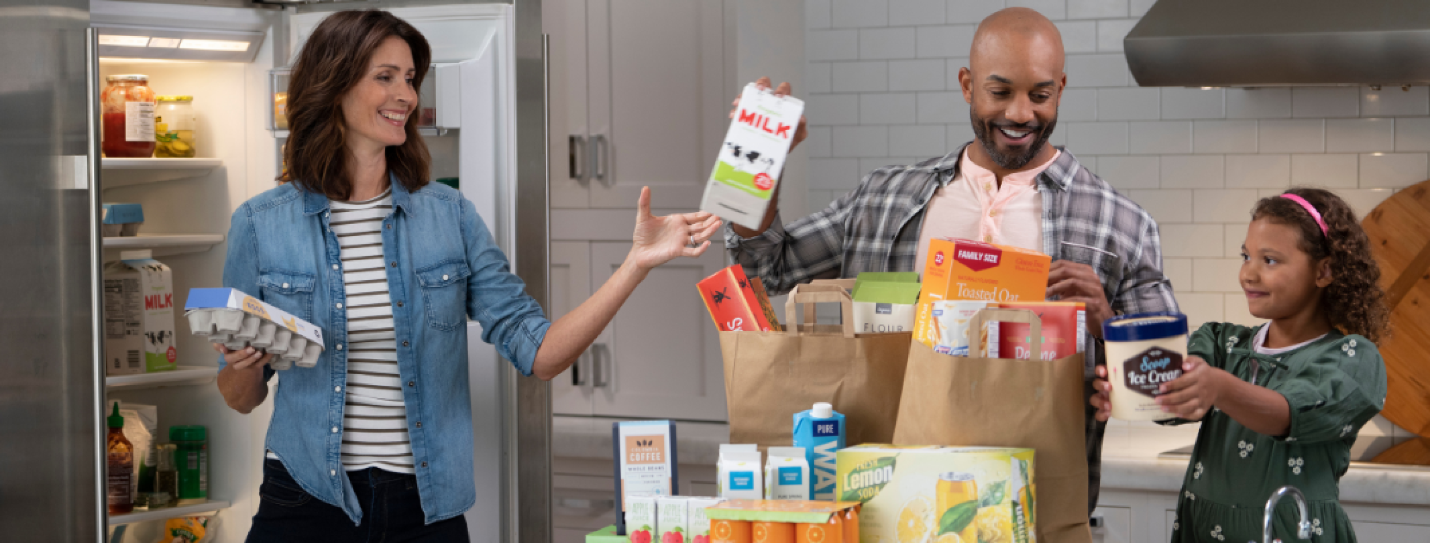 family unpacking their groceries. father is passing the mother a milk in paper carton to put in the fridge.