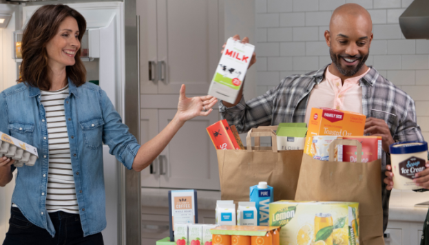 family unpacking their groceries. father is passing the mother a milk in paper carton to put in the fridge.